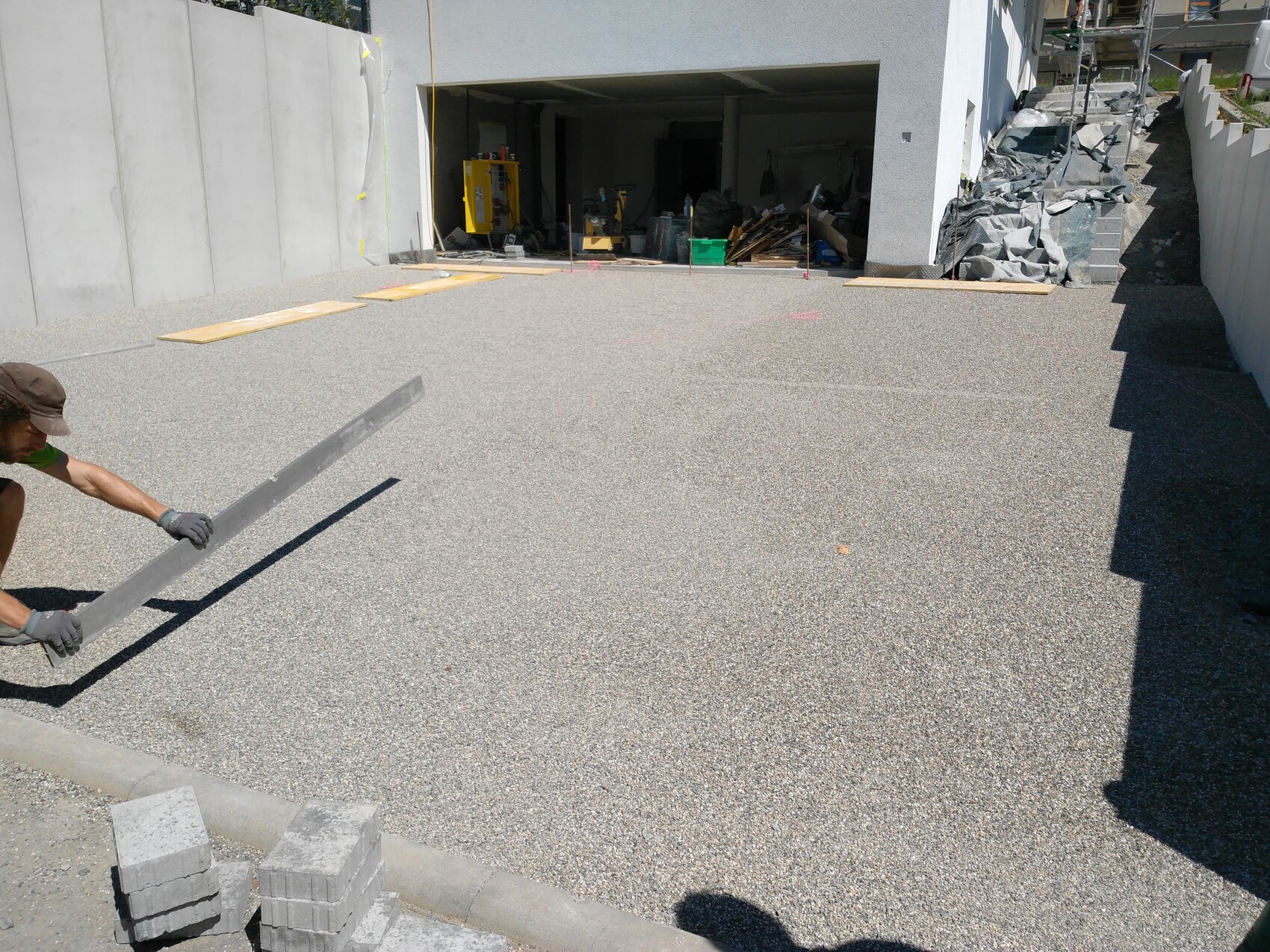 Bauarbeiter legt Metallleiste auf Kiesboden vor Garage, Baustellenmaterial sichtbar.