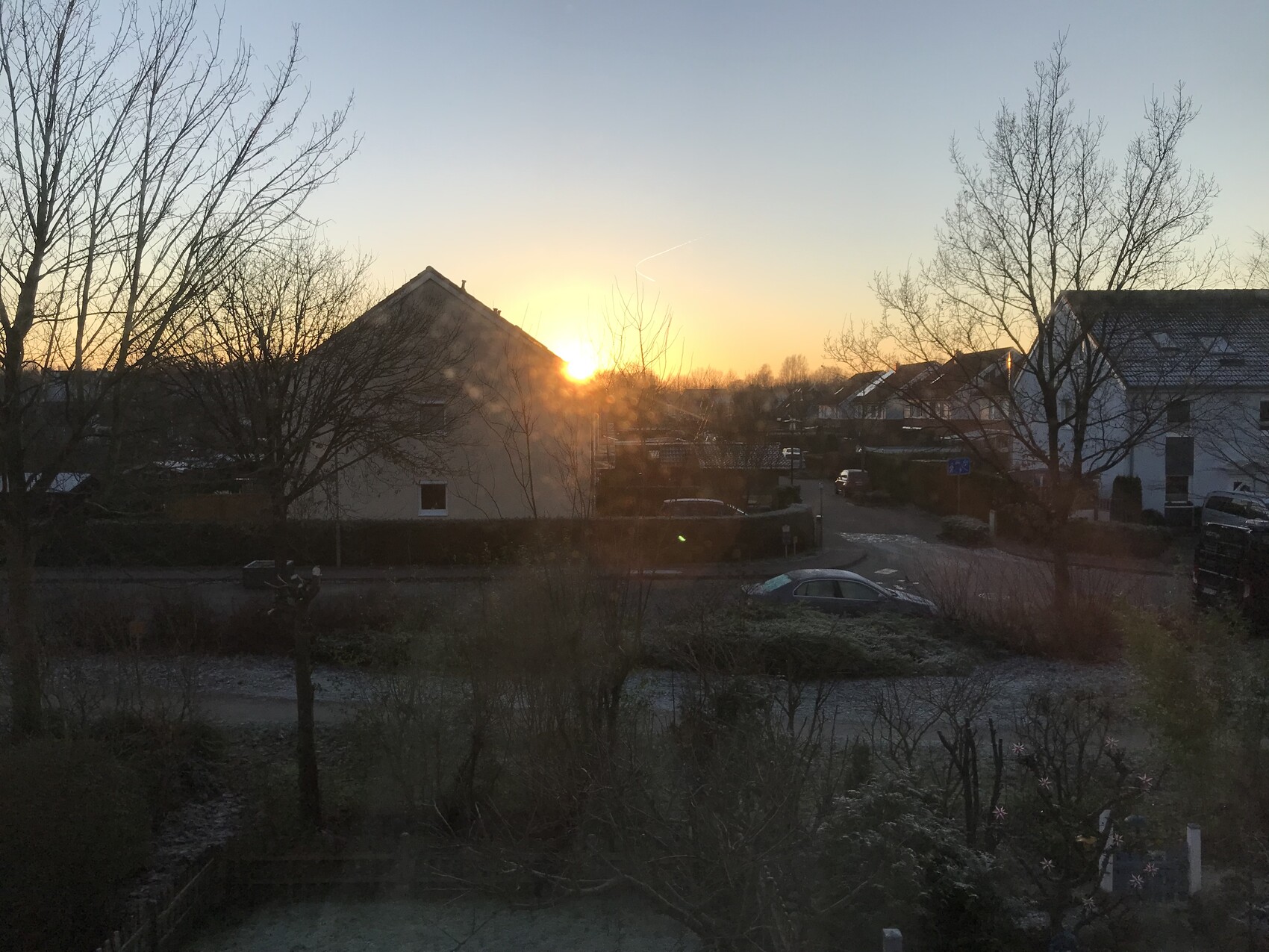 Abendsonne hinter Wohnhäusern in ruhiger Vorstadt, blattlose Bäume, geparkte Autos.