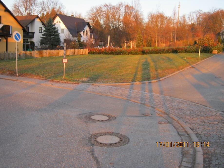 Wohngebiet mit Grasstreifen, Häuser links, Straßenecke, runde Kanaldeckel, lange Schatten