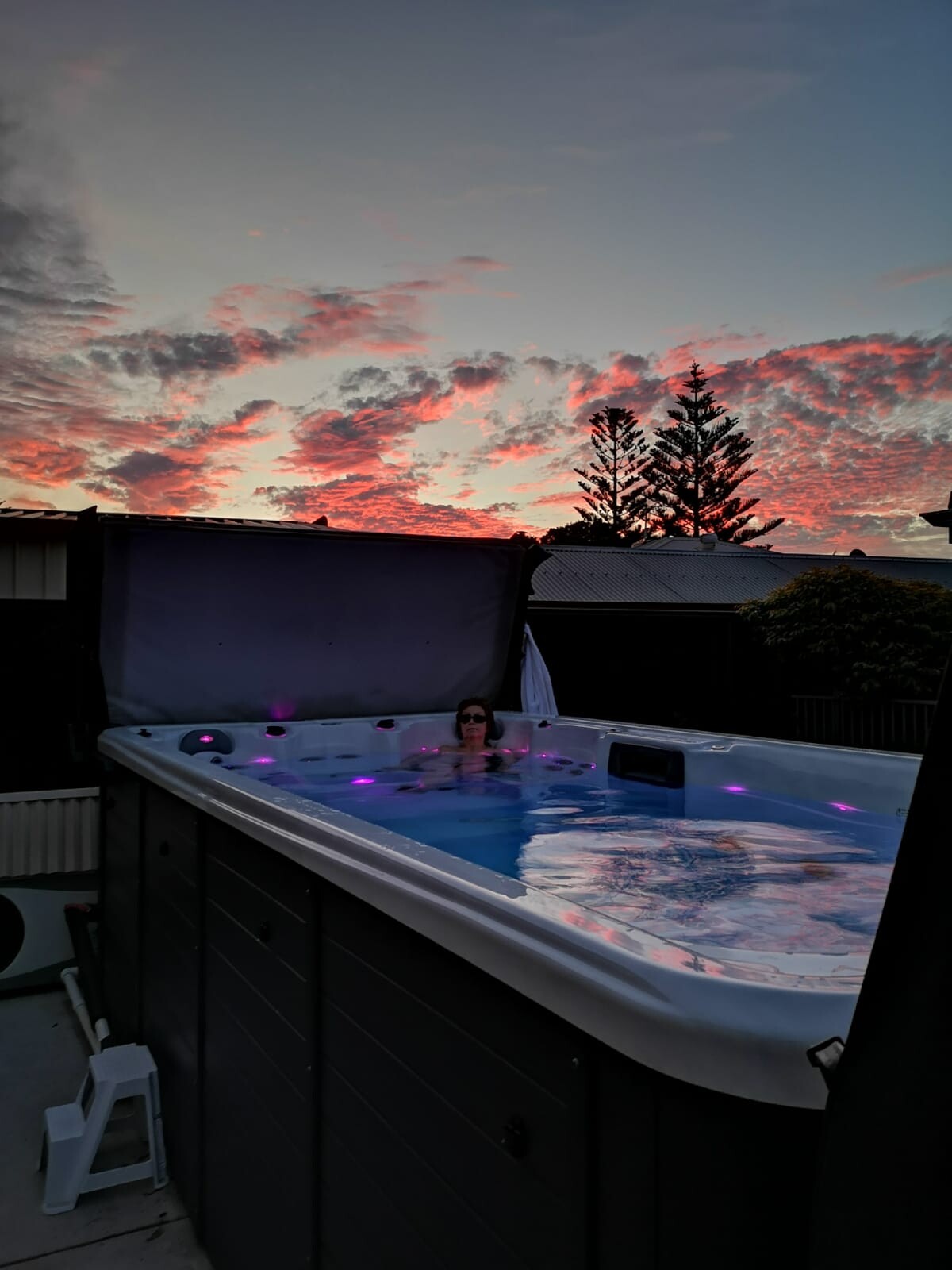 Person entspannt in einem beleuchteten Outdoor-Whirlpool mit violettem Licht bei Sonnenuntergang.