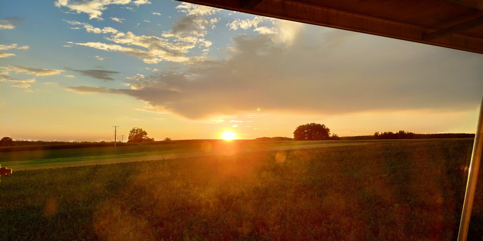 Sonnenuntergang über weiten Feldern, Blick von einer Terrasse auf offene Landschaft mit Bäumen.
