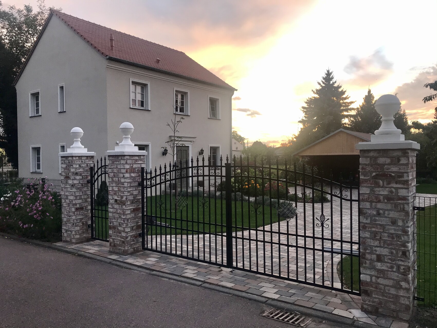 Helles, zweistöckiges Haus mit rotem Dach hinter schmiedeeisener Toreinfahrt; Garten, Sonnenuntergang.
