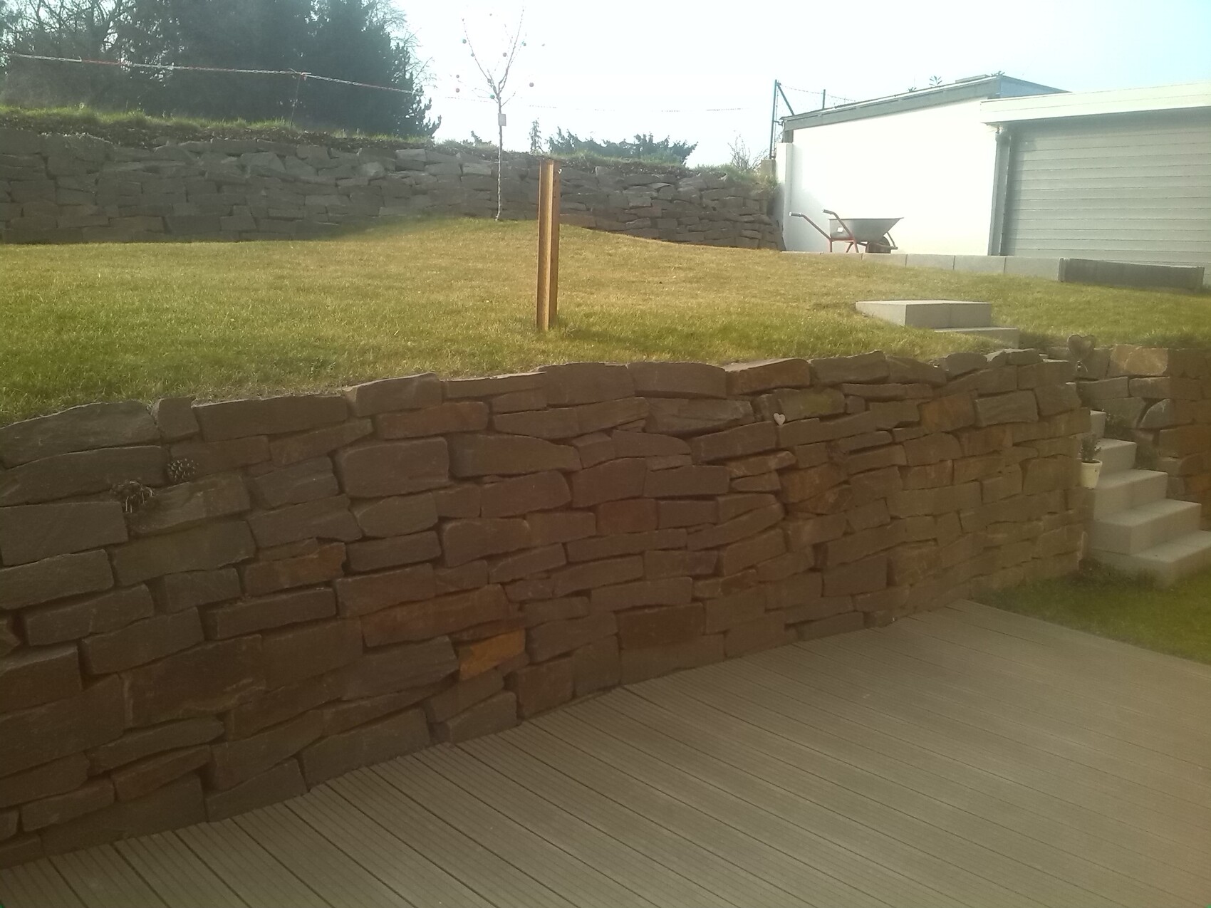 Trockenmauer aus Bruchsteinen vor einer Grasfläche; Treppenstufen rechts und Garage im Hintergrund.