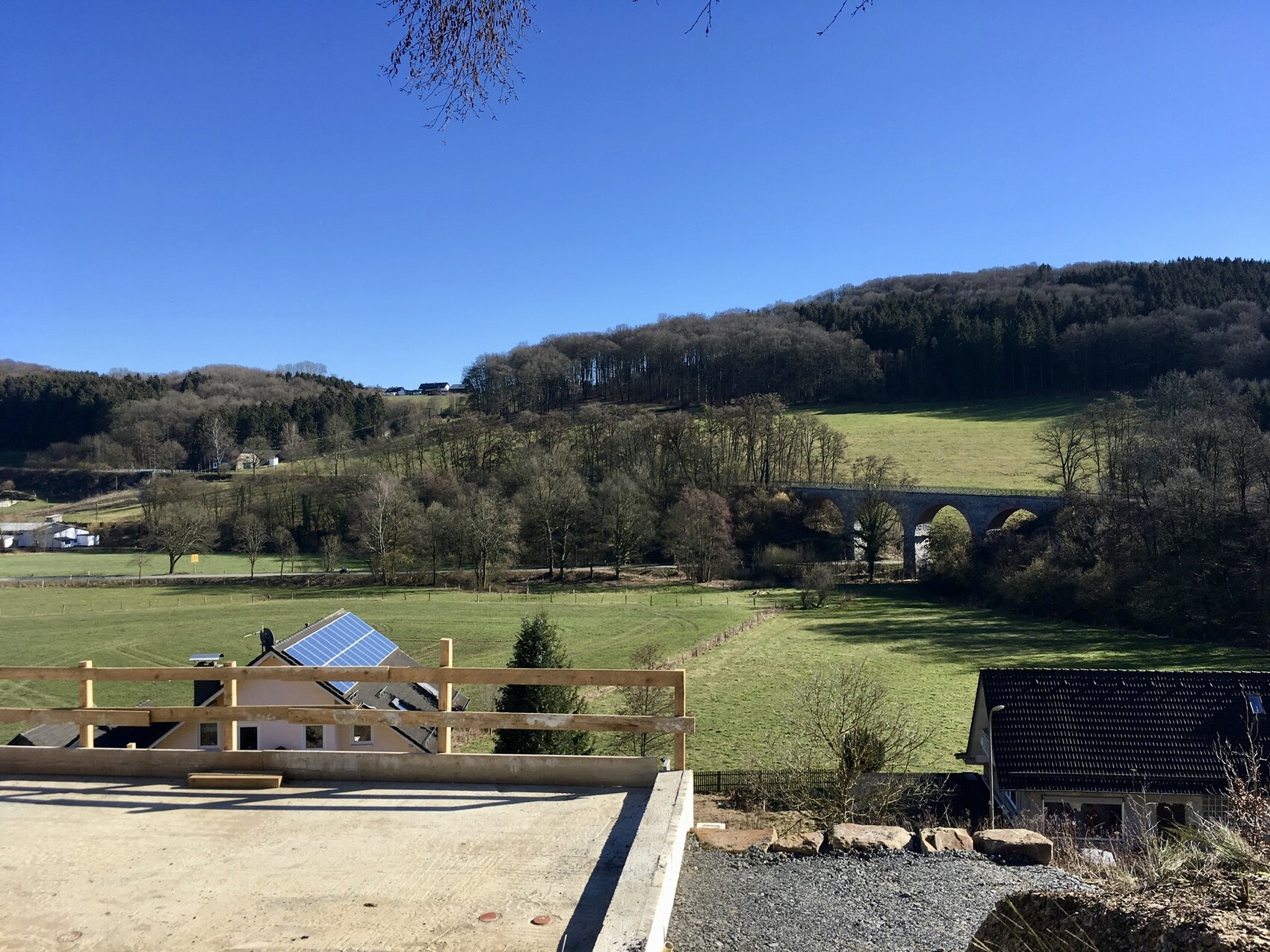 Blick über grüne Felder zum Steinbogenviadukt; Vordergrund Terrasse mit Solarpanelen.