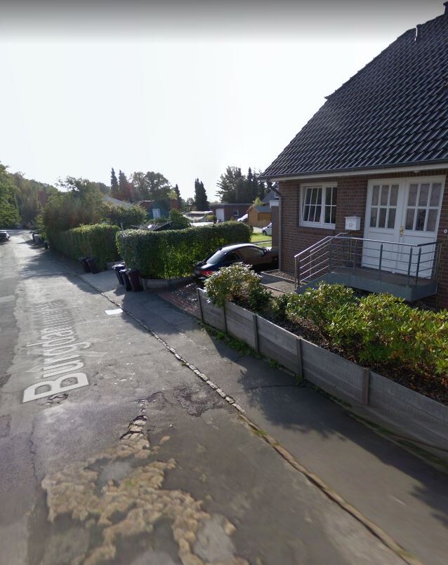 Straße im Vorort: braunes Backstein-Haus rechts, geparktes Auto, Hecken und Gartenzaun.