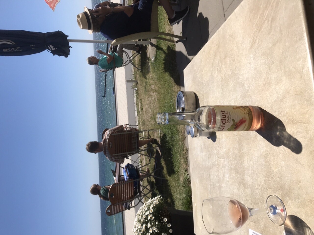 Terrasse am Meer bei sonnigem Wetter; Gruppe sitzt, Flasche und Gläser stehen auf dem Tisch.