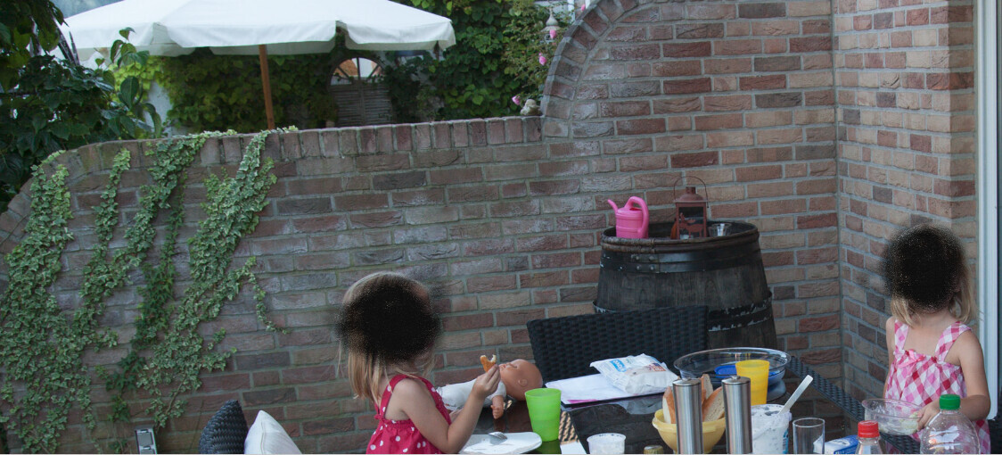 Zwei Mädchen essen draußen an einem Tisch; Spielzeugbaby liegt auf dem Tisch.