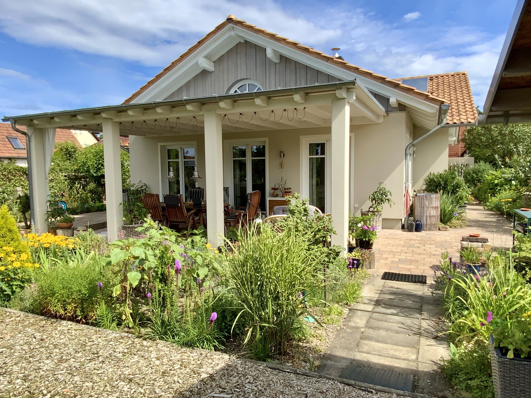 Einfamilienhaus mit überdachter Terrasse, Sitzgruppe unter Dach, bunte Gartenblumen.
