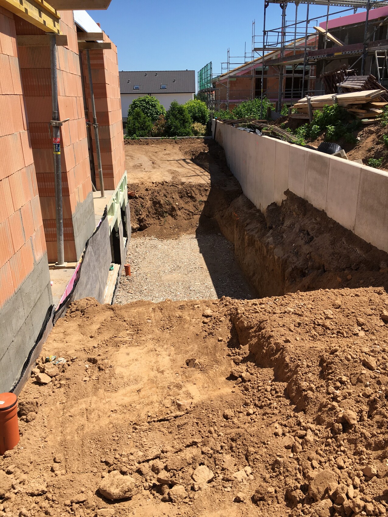 Baugrube neben unfertigem Ziegelhaus links, Betonmauer rechts, Gerüst und Holzstapel im Hintergrund.