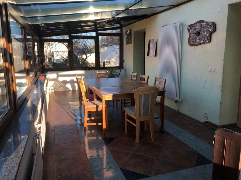 Heller Glas-Wintergarten mit großem Holztisch und Stühlen, Sonnenlicht durch Fenster von links.
