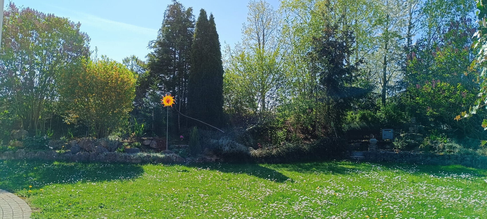 Garten mit grünem Rasen, Bäumen und Sträuchern; gelbe Sonnenblumen-Skulptur auf Stab.