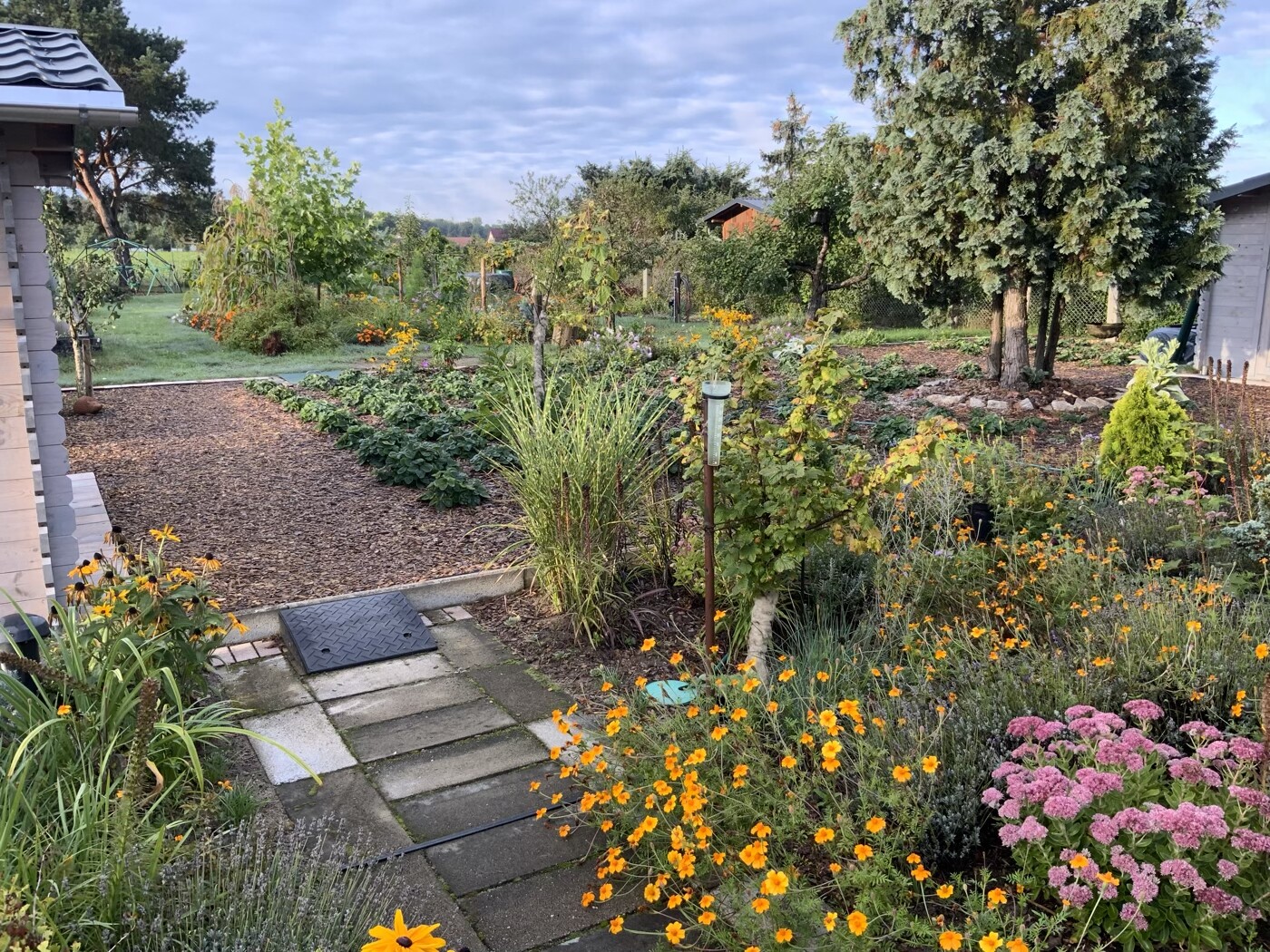 Garten mit Steinpfad, bunten Blumenbeete (orange, rosa), Bäume im Hintergrund.
