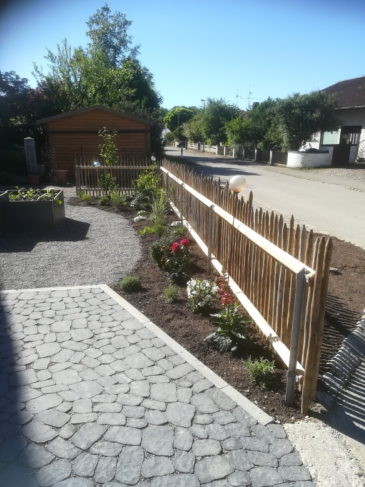 Garten mit Beet voller Blumen, Holzpalisaden-Zaun am Straßenrand, Holzhäuschen im Hintergrund