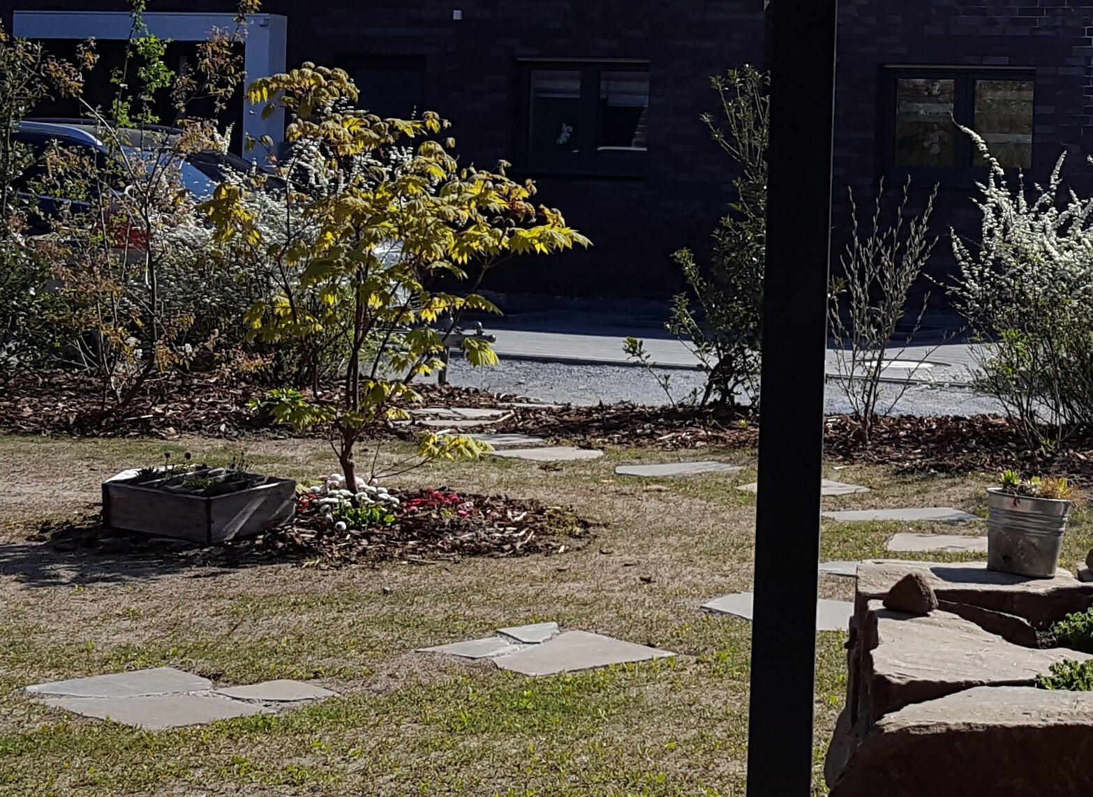 Garten mit kleinem Baum, Blumenbeet, Steinpfad, dunkler Pfosten im Vordergrund, Haus im Hintergrund