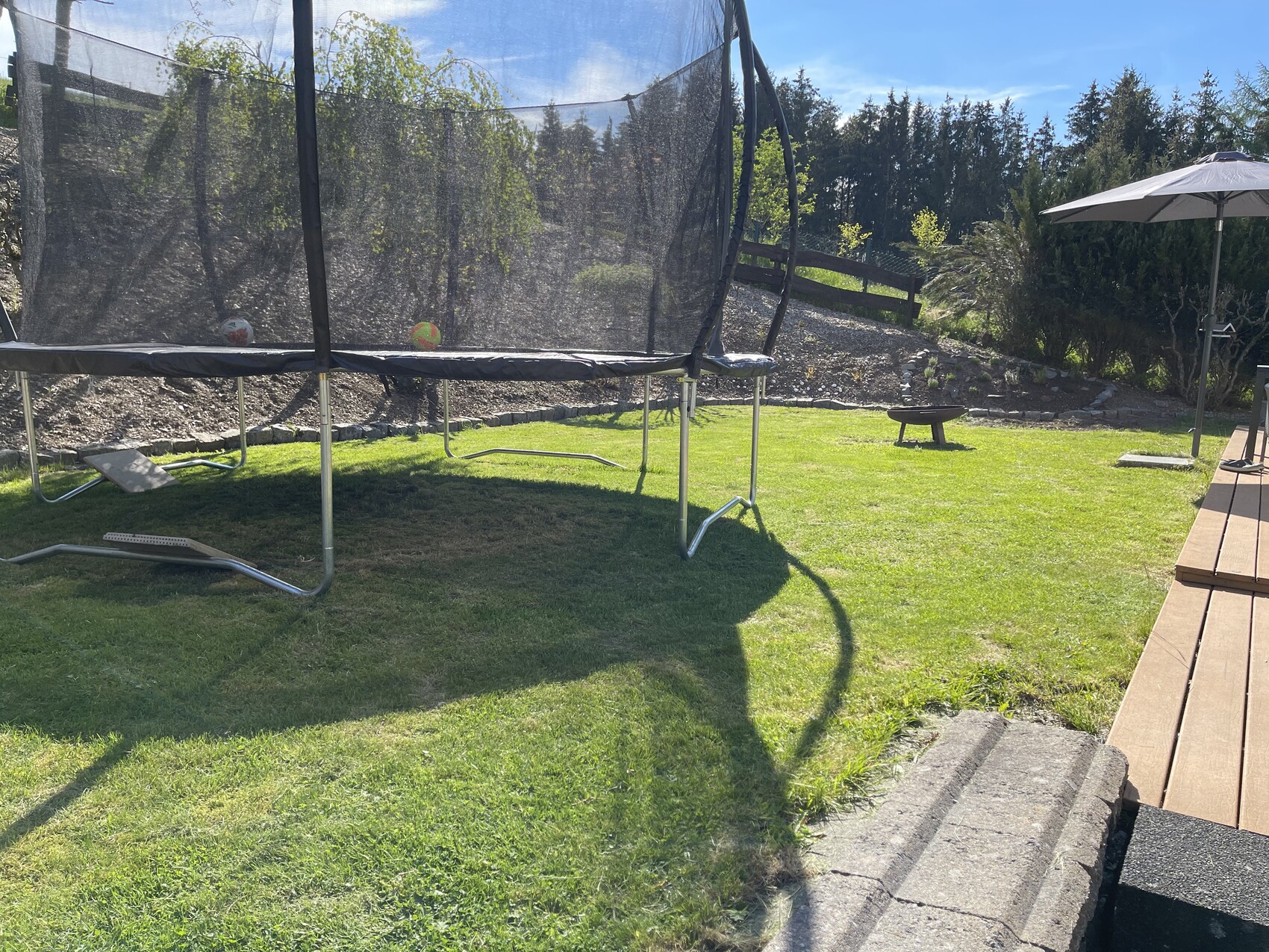 Großes Trampolin mit Sicherheitsnetz auf grünem Rasen, daneben Holzterrasse mit Sonnenschirm.