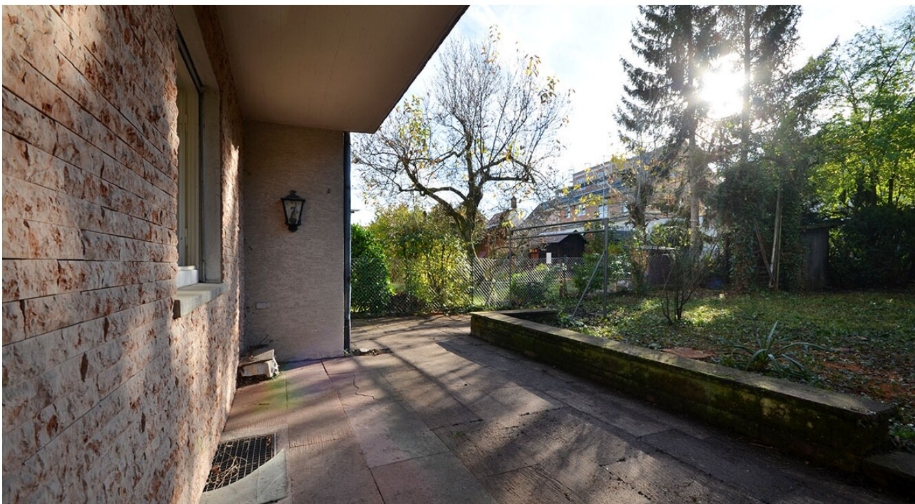 Außenansicht eines Hauses: Steinwand, Terrasse und Garten mit Zaun, Sonnenlicht.