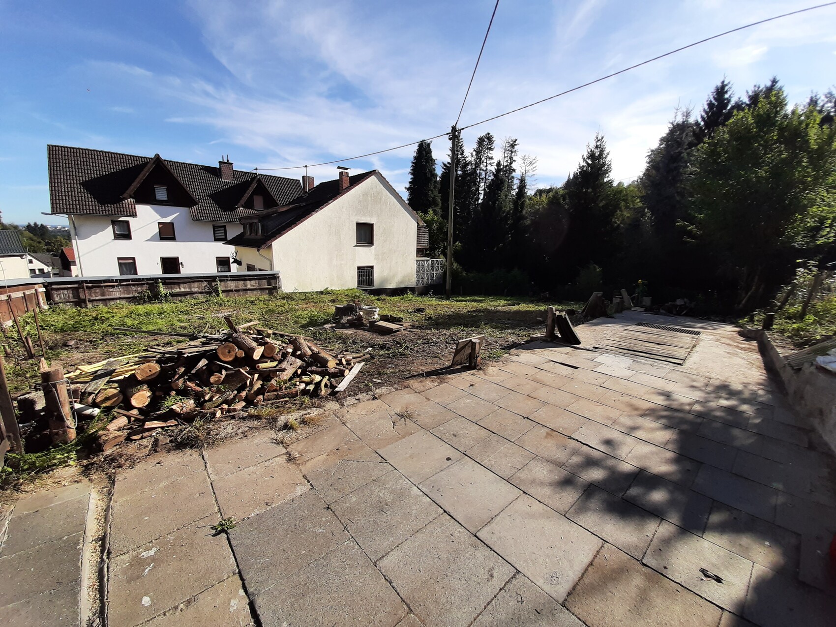 Gepflasterter Hof mit Holzstapel, weißes Haus dahinter, Baumreihe rechts.