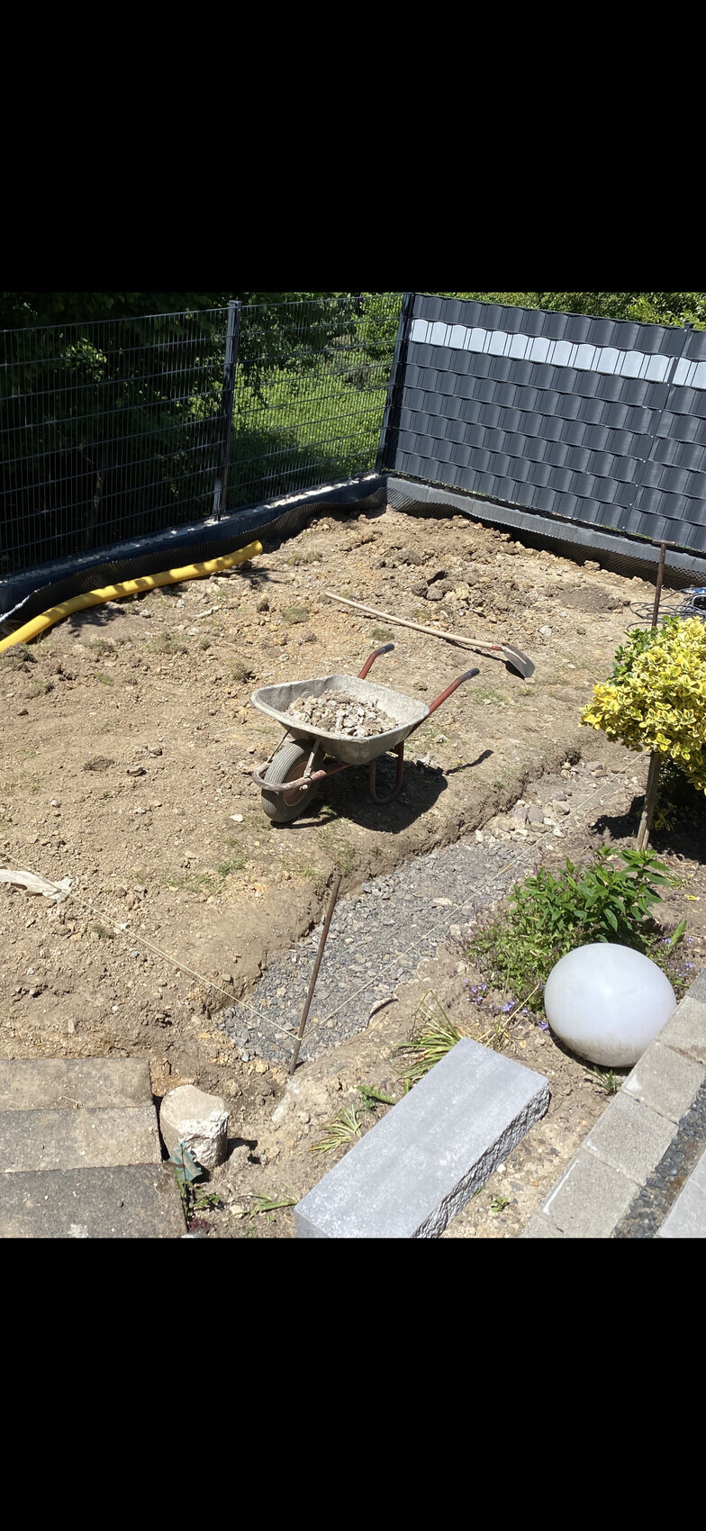 Gartenbaustelle: Schubkarre mit Kies, Spaten und Schaufel, lose Erde, Zaun im Hintergrund.