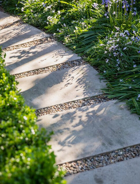Gartenpfad mit flachen Steinplatten, Kies dazwischen und Rand aus Gras und lila Blumen.