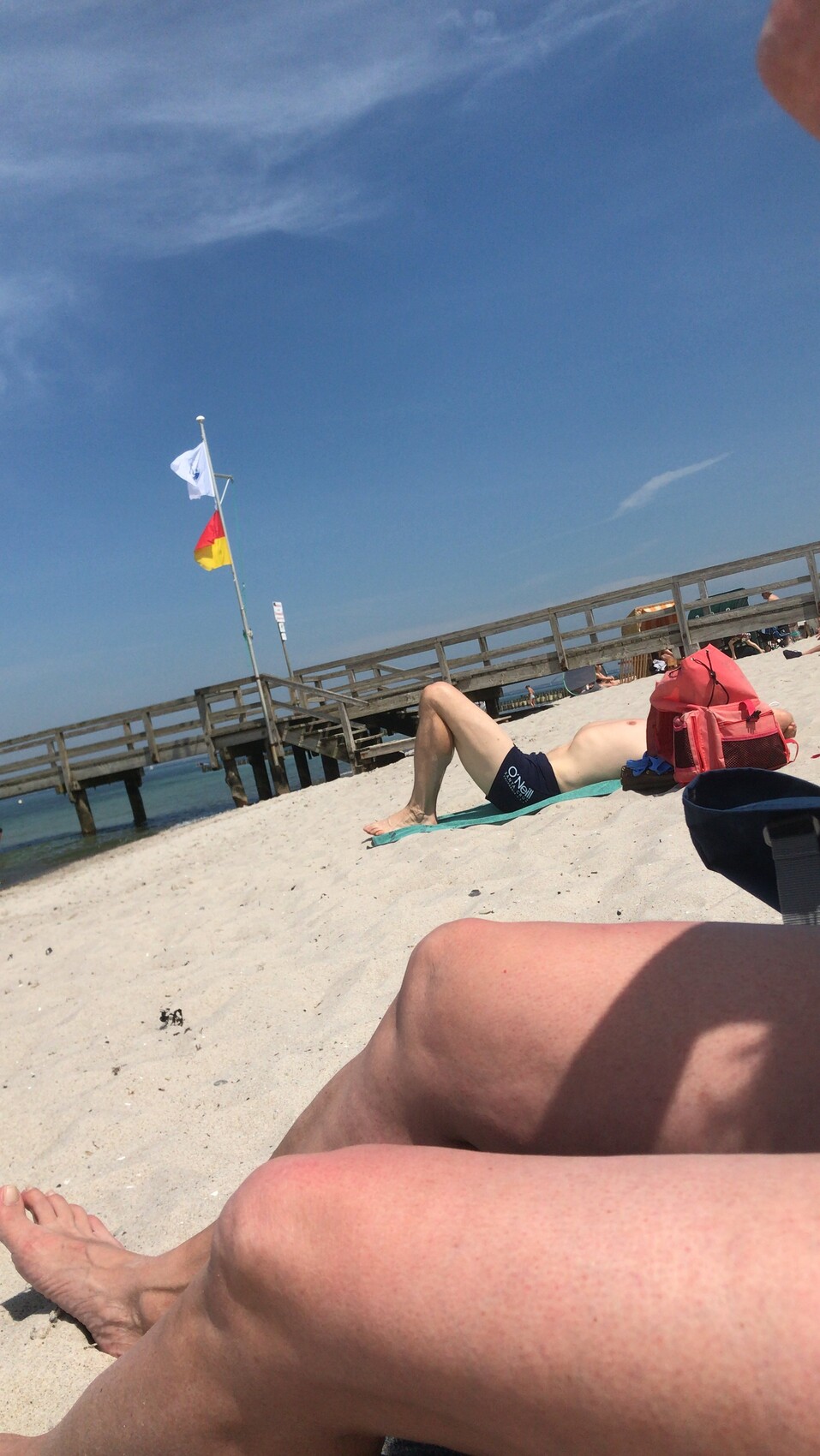 Person liegt entspannt am Strand neben roter Tasche; Holzsteg und Flaggen im Hintergrund.