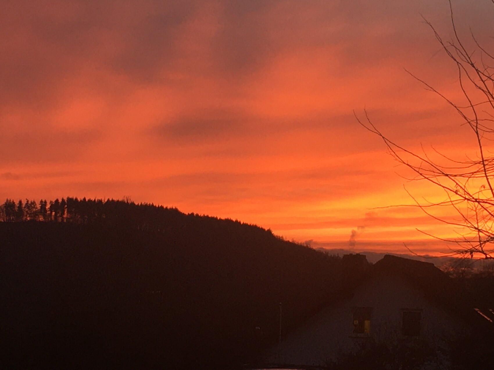 Oranger Sonnenuntergang, dunkle Silhouette von Hügeln, Wald und Häusern; rechts kahle Äste.
