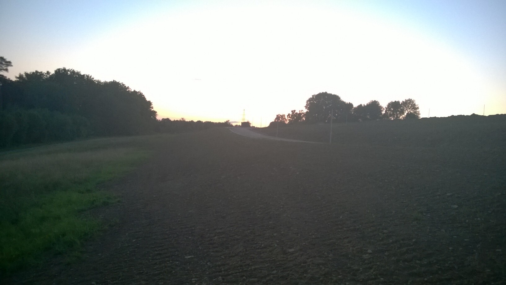 Landschaft mit Schotterweg durch ein Feld, Bäume am Rand, Sonnenuntergang am Horizont