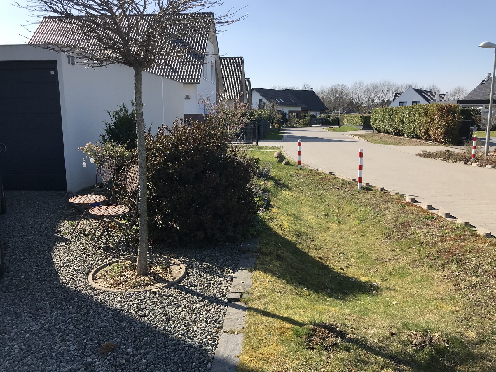 Vorortblick mit Baum, Metallstühle, Kiesfläche, Hecke und Straße mit Pfosten.