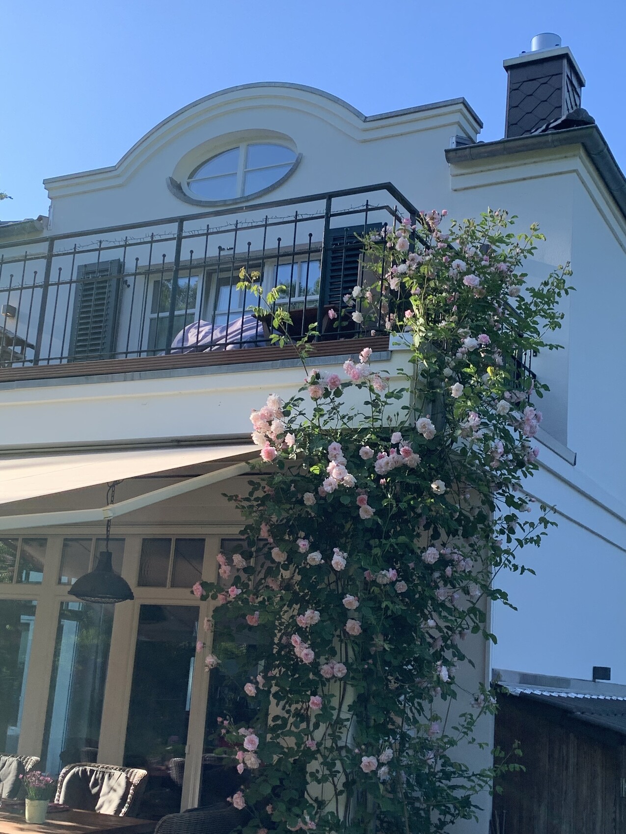 Weiße Hausfassade mit Balkon und rundem Oberfenster, rosa Rosen an der Wand; Terrasse mit Möbeln.