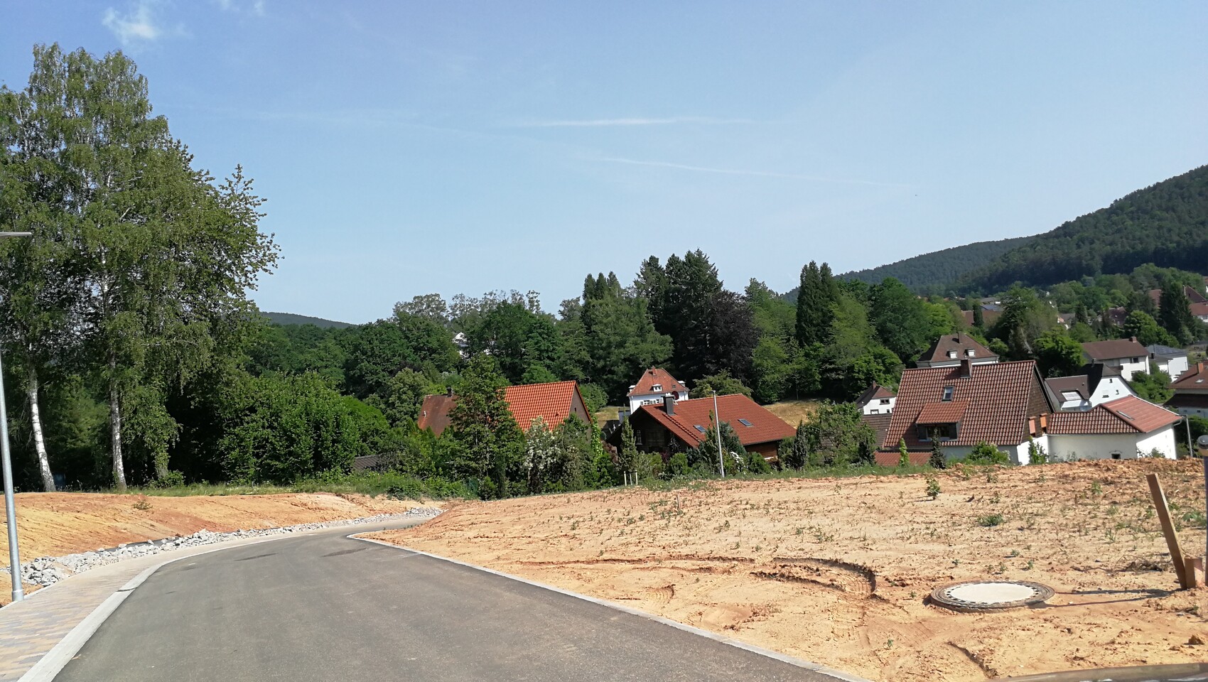 Straße im Bau, Sandboden, Häuser mit roten Ziegeldächern, Bäume und Hügel dahinter.