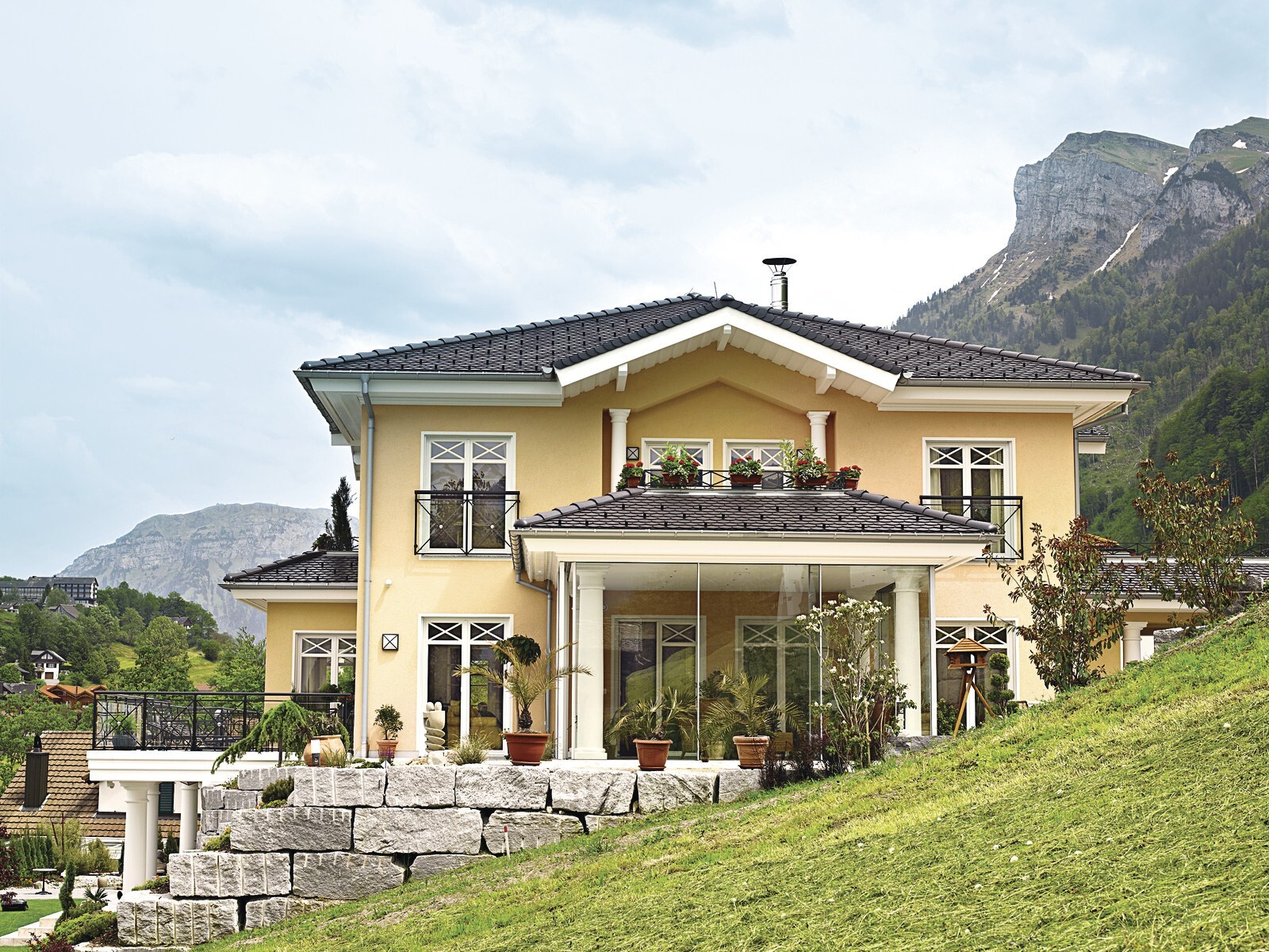 Gelbes Einfamilienhaus auf Hang mit Balkon, Glasveranda und Berglandschaft im Hintergrund