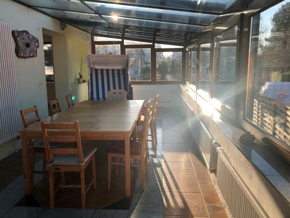 Sonnendurchflutete Veranda mit großem Holztisch, Stühlen und Glasdach, Blick nach außen.