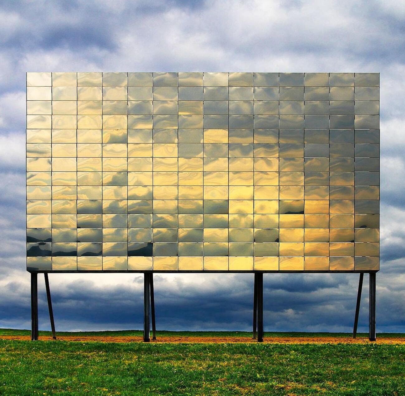 Großes rechteckiges Spiegelpaneel aus Metallplatten auf Beinen vor Wolken.