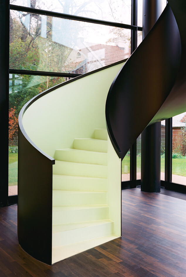 Spiraltreppe im Innenraum neben grossen Glasfenstern, dunklem Boden und schwarzer Saeule.
