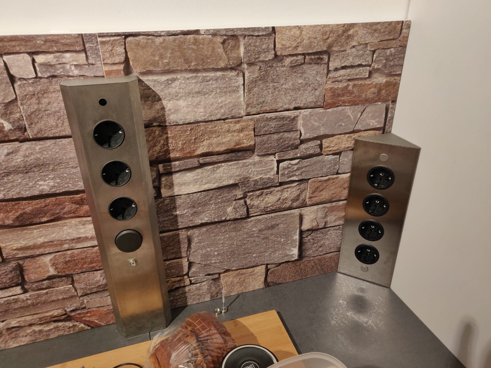 Zwei Edelstahl-Steckdosenleisten vor Steinwand in der Küche mit Brot auf dem Tisch