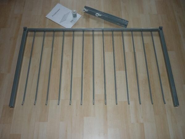 Metallrahmen mit senkrechten Stäben liegt auf Holzboden; Anleitung und Montagematerial daneben.