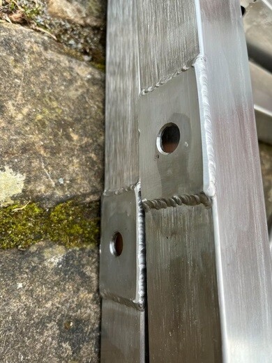 Nahaufnahme eines Metallrahmens mit Schweißnähten und Bohrungen neben Steinboden