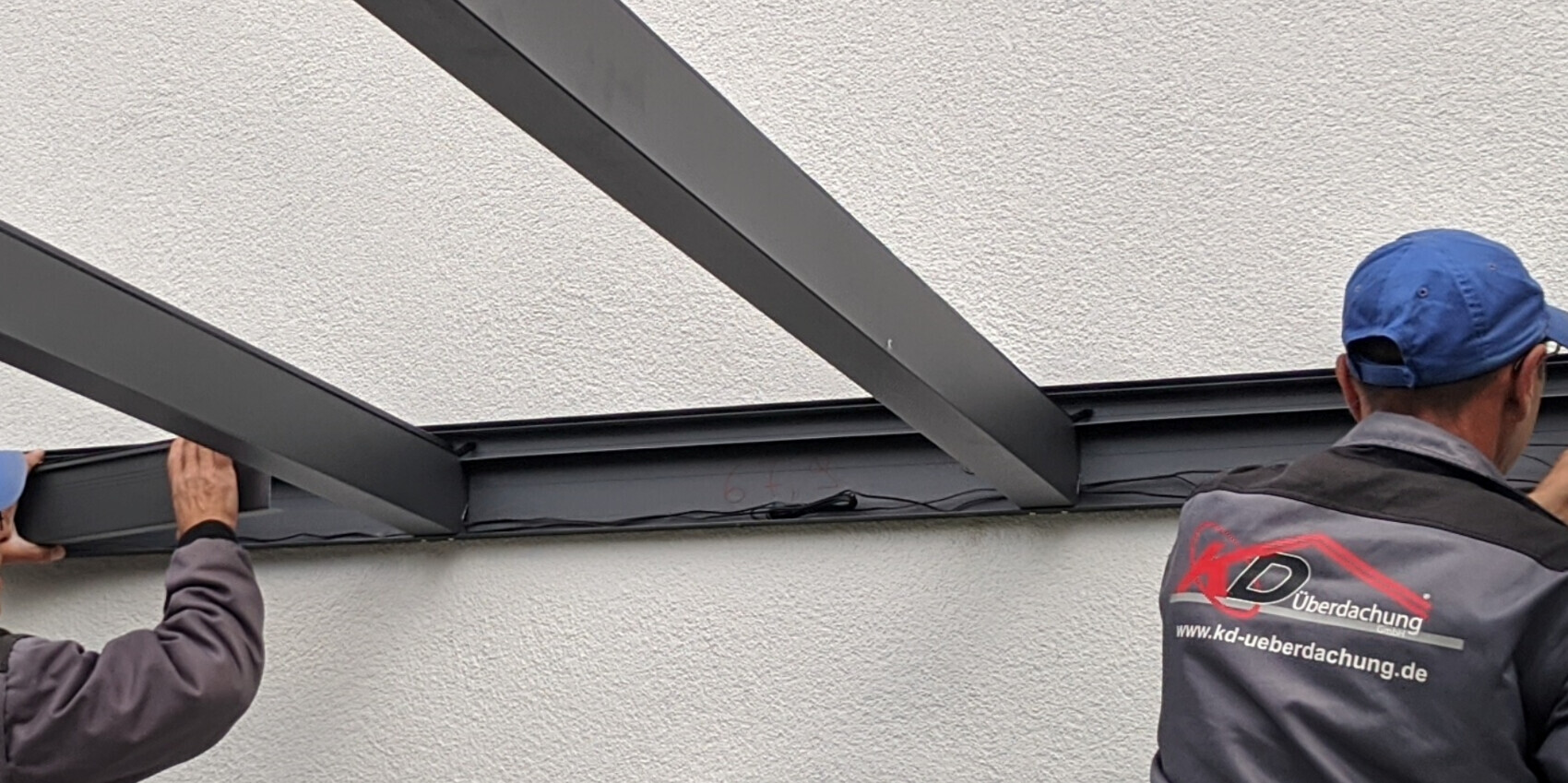 Zwei Arbeiter montieren dunkle Stahlträger an einer weißen Außenwand zu einer Überdachung.