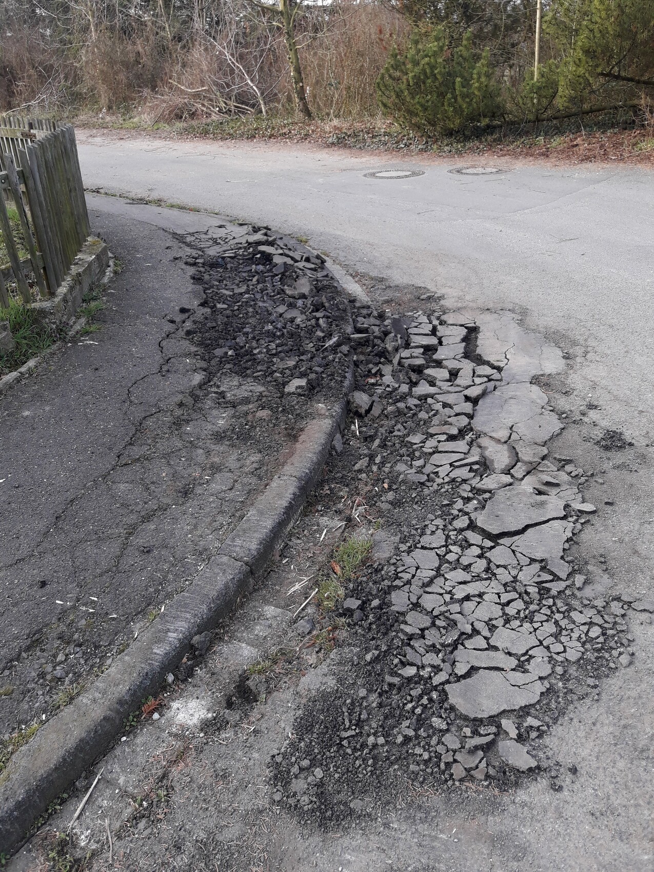 Abgebrochene Asphaltdecke am Straßenrand; Geröll, Bruchstücke, links Holzzaun.