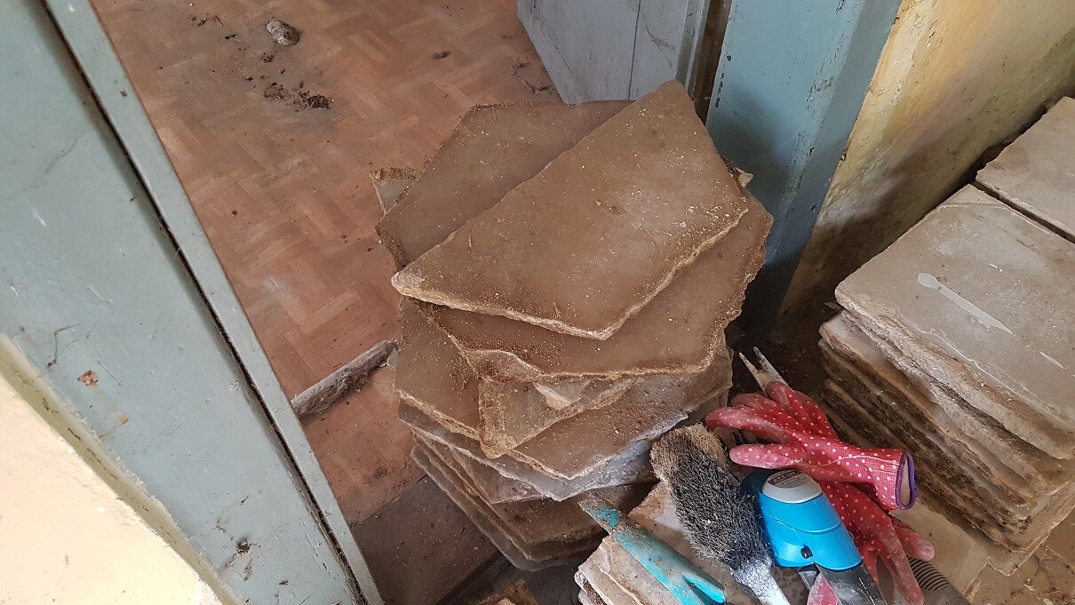 Staubige Baustelle mit gestapelten braunen Bodenbelägen, Reinigungswerkzeug und Handschuhen.