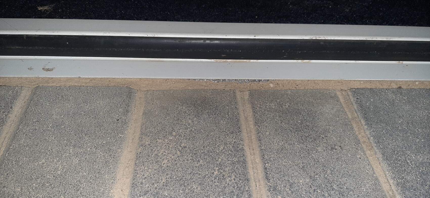Türschwelle mit grauem Metallrahmen, schwarzem Bodenbelag und staubigen Betonplatten.