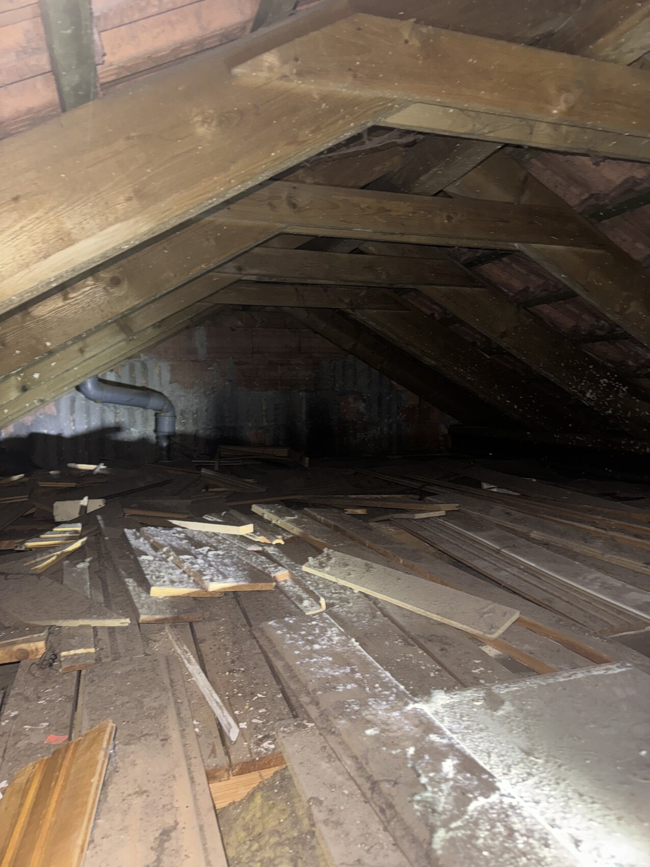 Dachboden mit sichtbarem Holzdachstuhl, verstreuten Brettern, Staub und dunkler Ecke im Hintergrund.