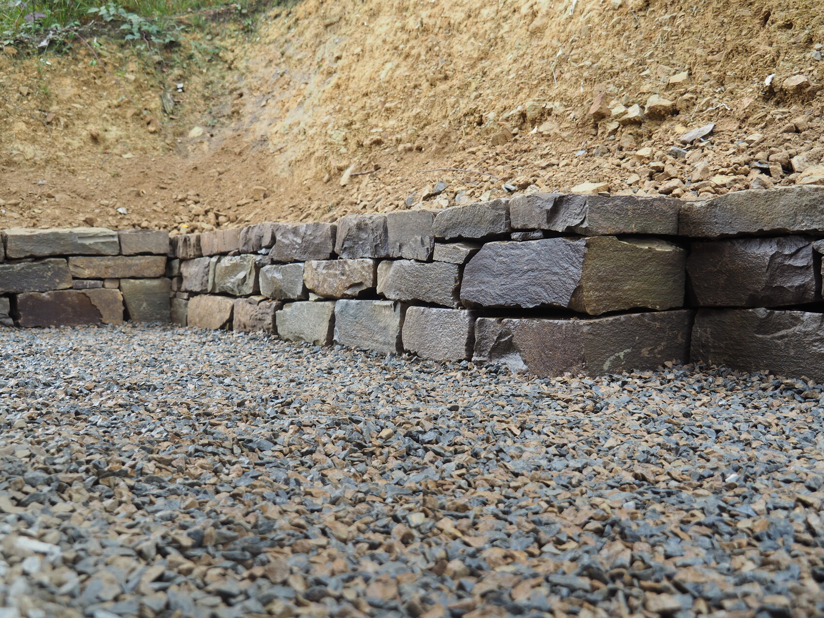 Stützmauer aus groben Natursteinen vor Kiesboden und Erdwall.
