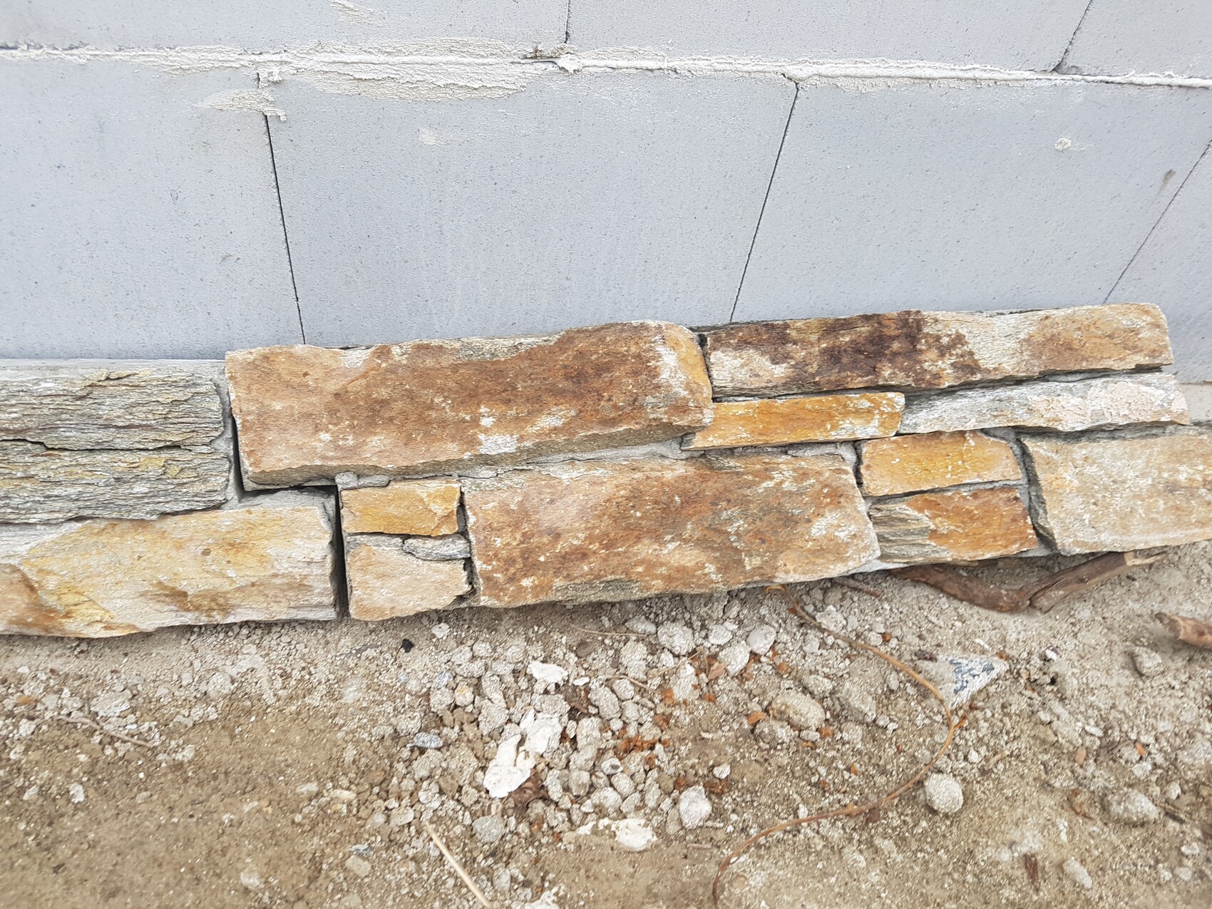 Niedrige Steinmauer aus unregelmäßigen braunen Natursteinen vor grauer Wand; Boden aus Erde und Kies.