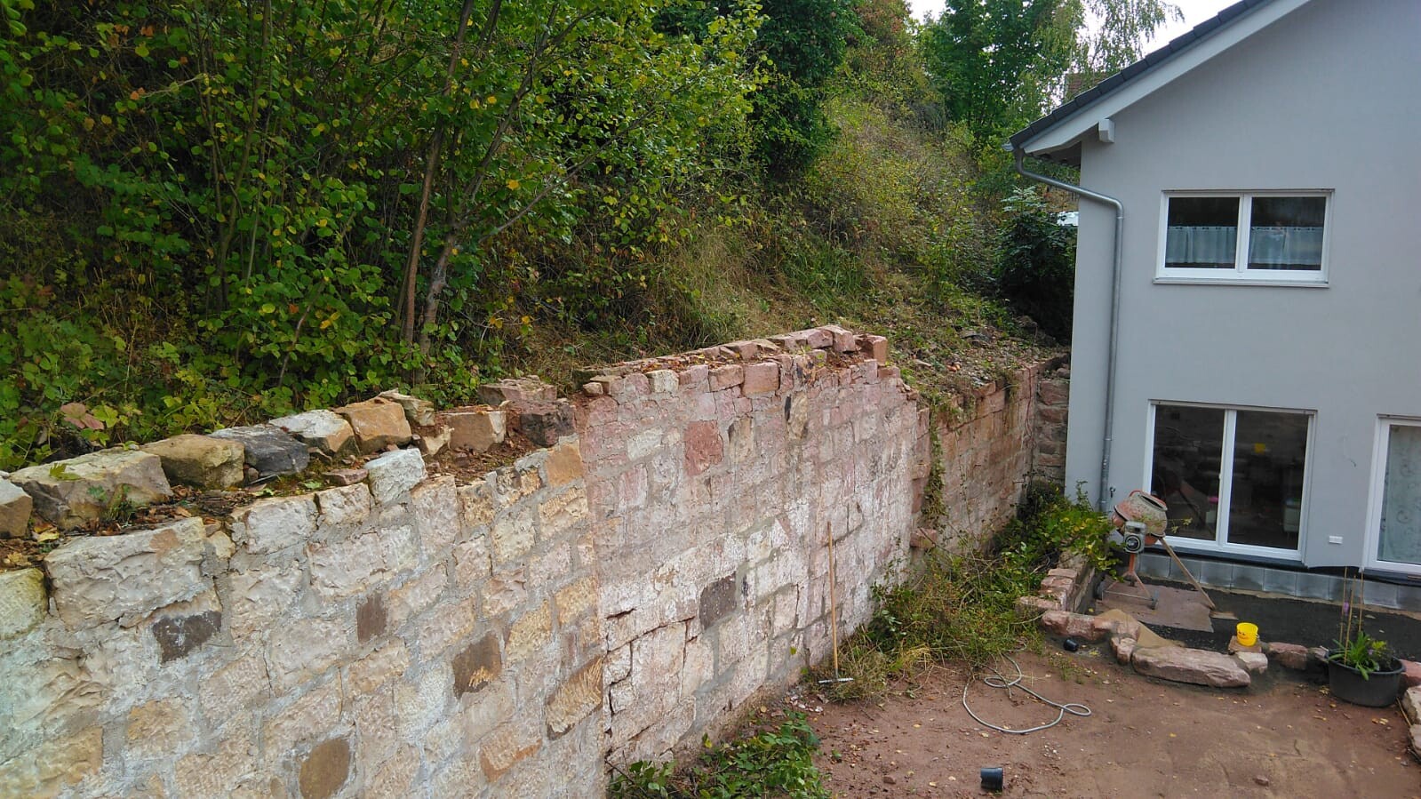 Außenansicht eines Hauses mit grauer Fassade, Fenstern und einer alten Steinmauer im Garten