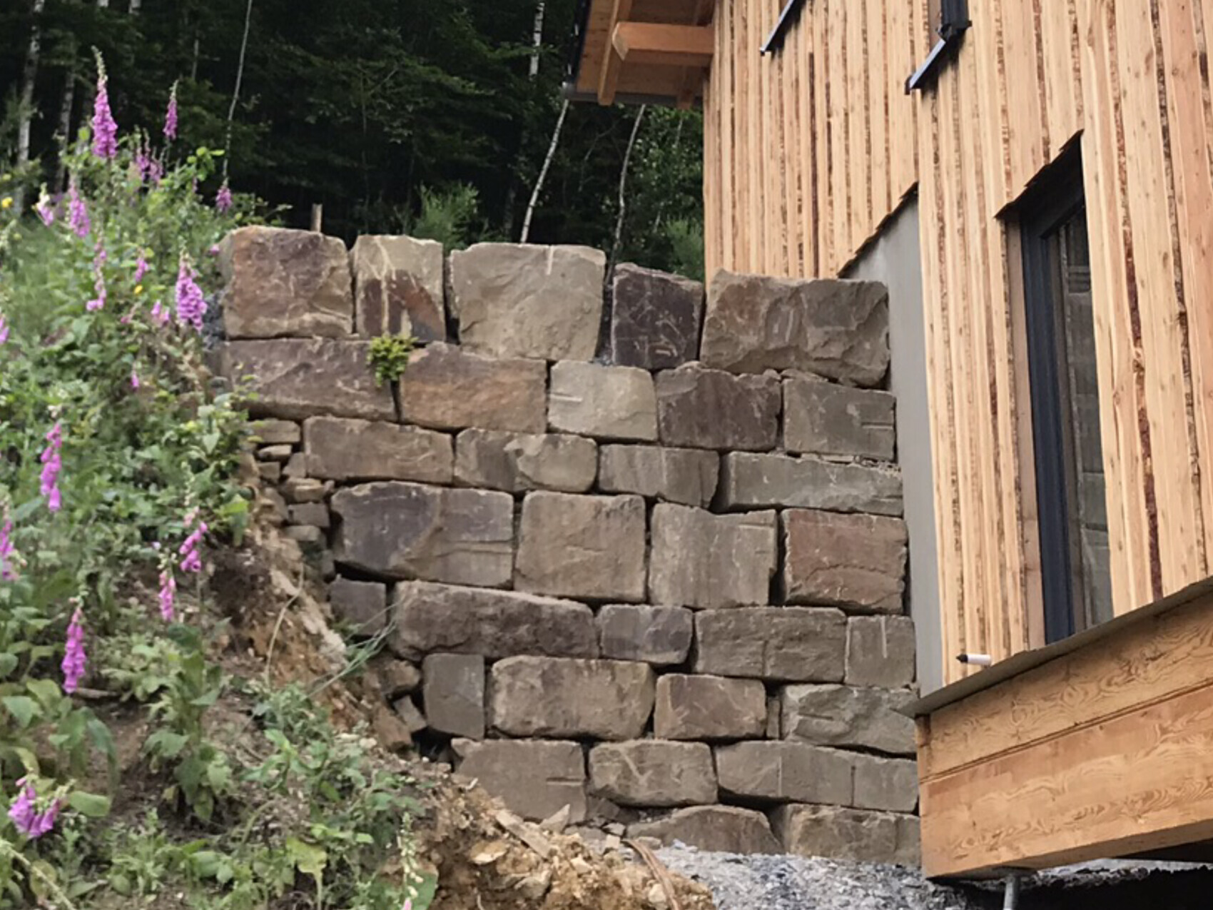 Große Natursteinmauer als Stützwand neben einem Holzhaus; linke Blumenpflanzen mit lila Blüten.