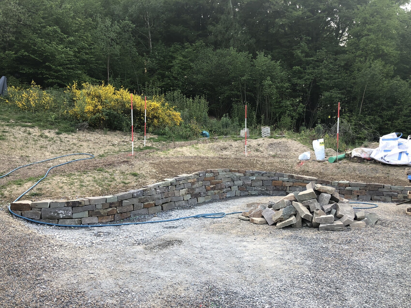 Kreisförmige Steinmauer aus Natursteinen, Baumaterialien und Schlauch im Baugelände.