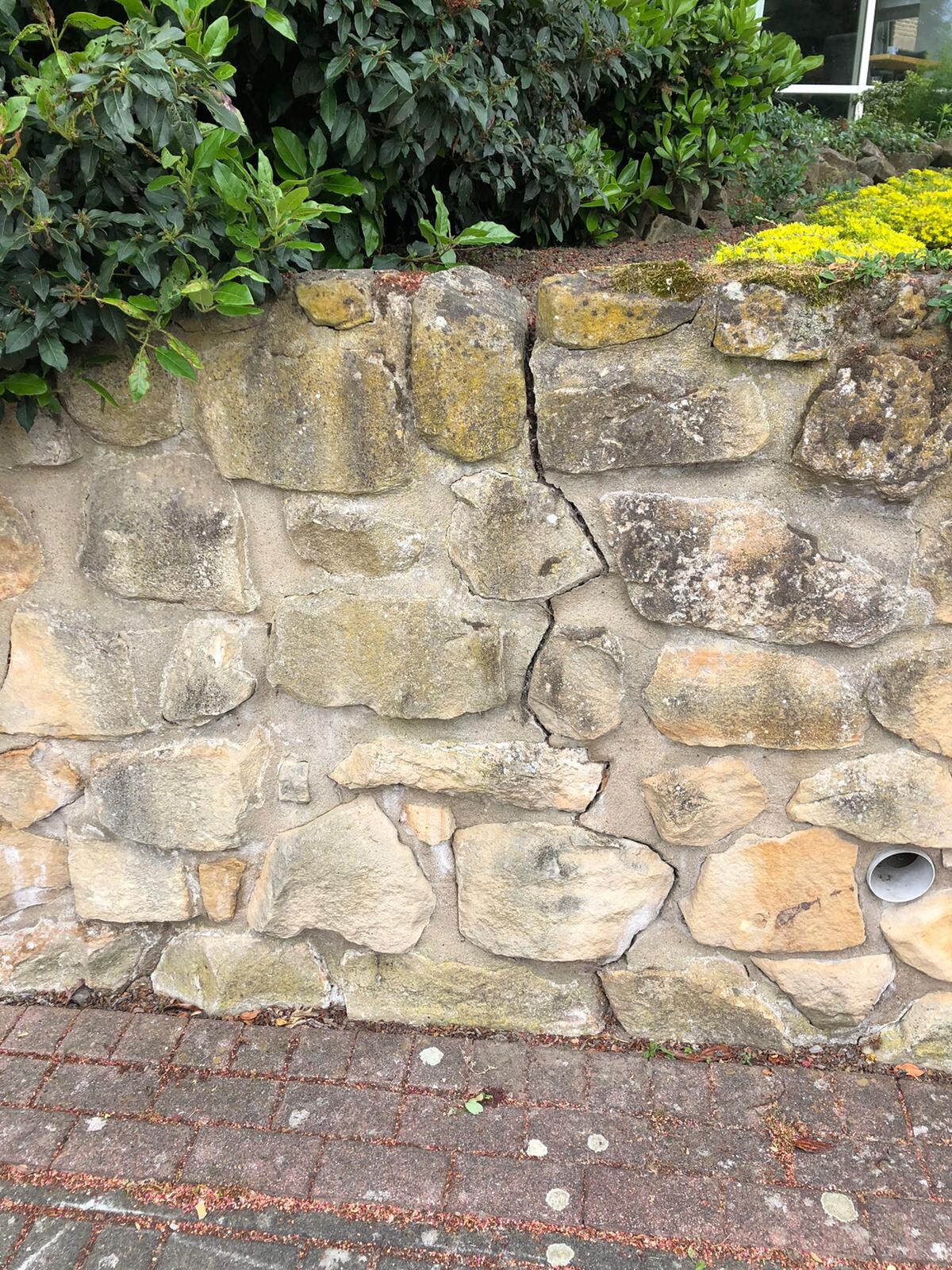 Steinmauer mit Riss in der Mitte, davor Gehweg aus Pflastersteinen, grüne Büsche oben.