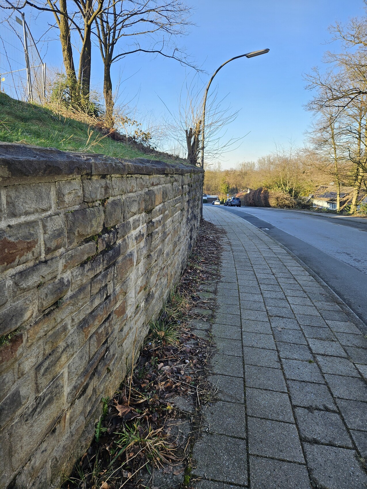 Gepflasterter Gehweg neben einer Steinmauer, gekrümmte Straßenlaterne, Autos im Hintergrund.