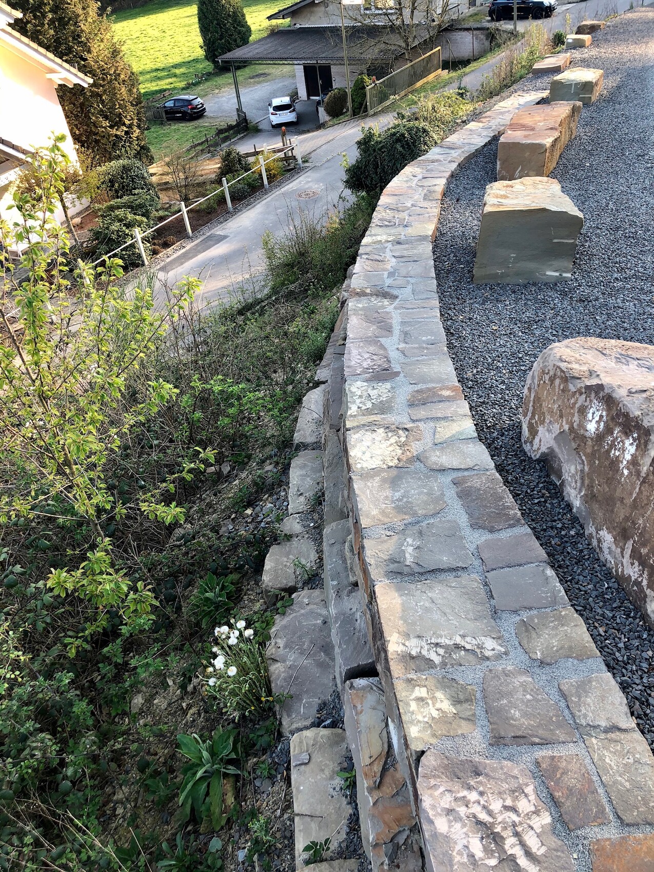 Steinmauer am Abhang mit Kiesfläche und großen Quadersteinen; Gartenpflanzen vorn.