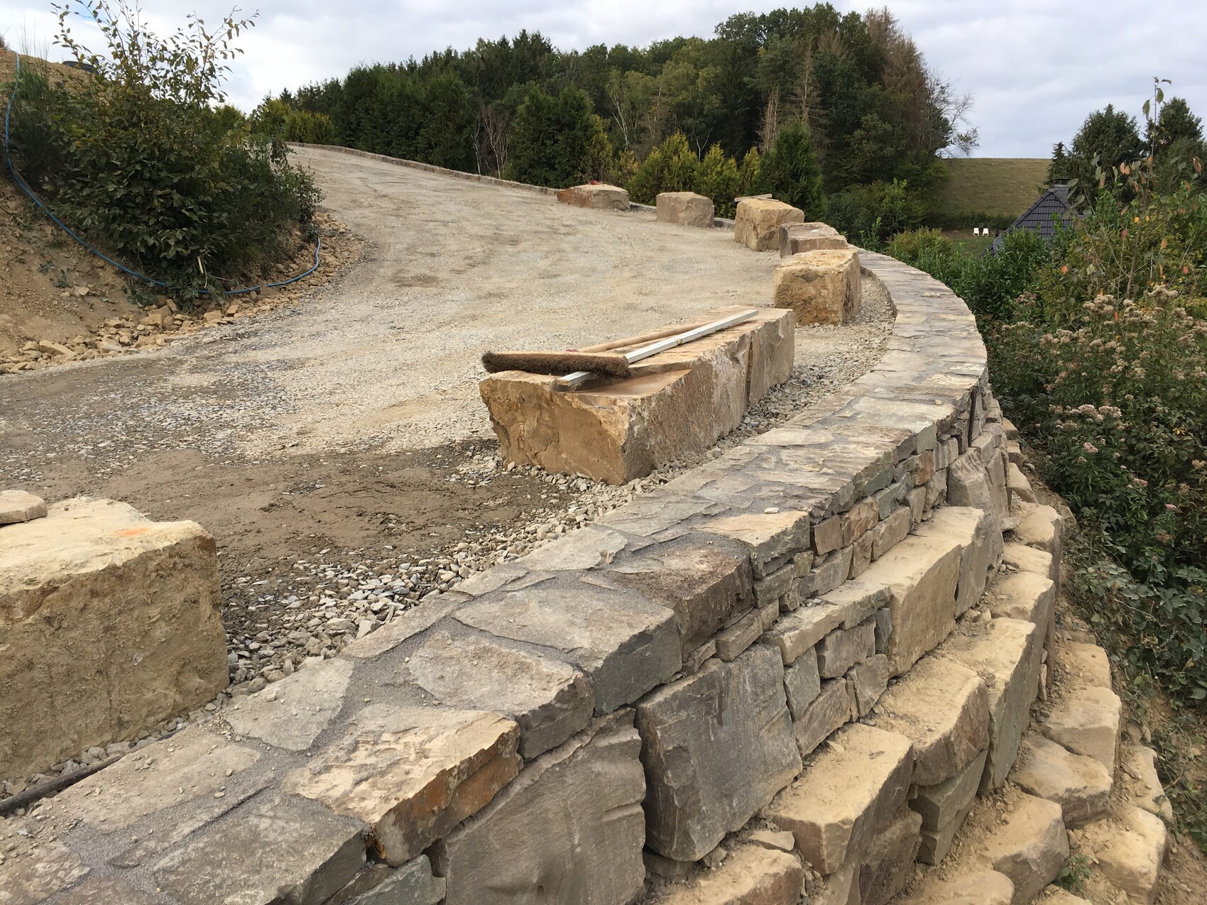 Natursteinmauer am Hang, unbefestigte Zufahrt, Kies und Steinblock mit Werkzeugen.